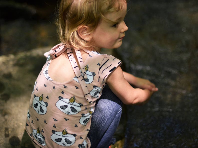 May include: A young girl wearing a pink and black panda print shirt with a criss-cross back detail. The shirt has a short sleeve and a relaxed fit.