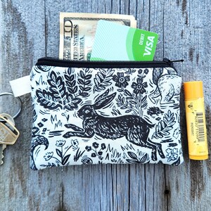 May include: A white zippered pouch with a black and white print of a rabbit, flowers, and leaves. The pouch is filled with cash, a credit card, and a lip balm.