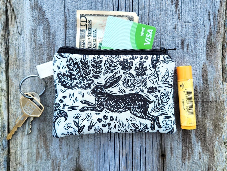 May include: A white zippered pouch with a black and white print of a rabbit, flowers, and leaves. The pouch is filled with cash, a credit card, and a lip balm.