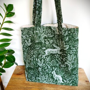 May include: Green tote bag with a white woodland pattern featuring rabbits, squirrels, and foliage. The bag has matching green handles and a cream-colored interior. The tote is made of fabric and is designed for carrying items.