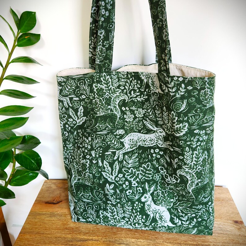 May include: Green tote bag with a white woodland pattern featuring rabbits, squirrels, and foliage. The bag has matching green handles and a cream-colored interior. The tote is made of fabric and is designed for carrying items.
