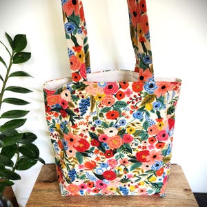 K&ouml;nnte beinhalten: Eine florale Tragetasche mit cremefarbenem Hintergrund und einem lebendigen Muster aus roten, rosa, blauen und gelben Blumen. Die Tasche hat zwei passende Griffe und ist aus robustem Stoff gefertigt. Die Tasche steht auf einer Holzoberfl&auml;che.