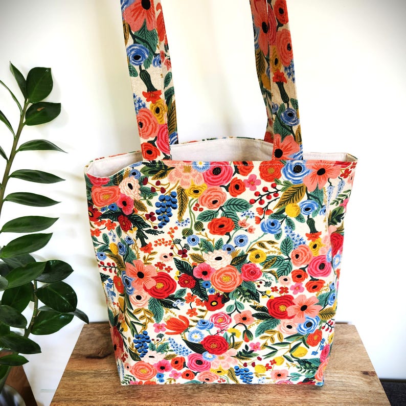 K&ouml;nnte beinhalten: Eine florale Tragetasche mit cremefarbenem Hintergrund und einem lebendigen Muster aus roten, rosa, blauen und gelben Blumen. Die Tasche hat zwei passende Griffe und ist aus robustem Stoff gefertigt. Die Tasche steht auf einer Holzoberfl&auml;che.