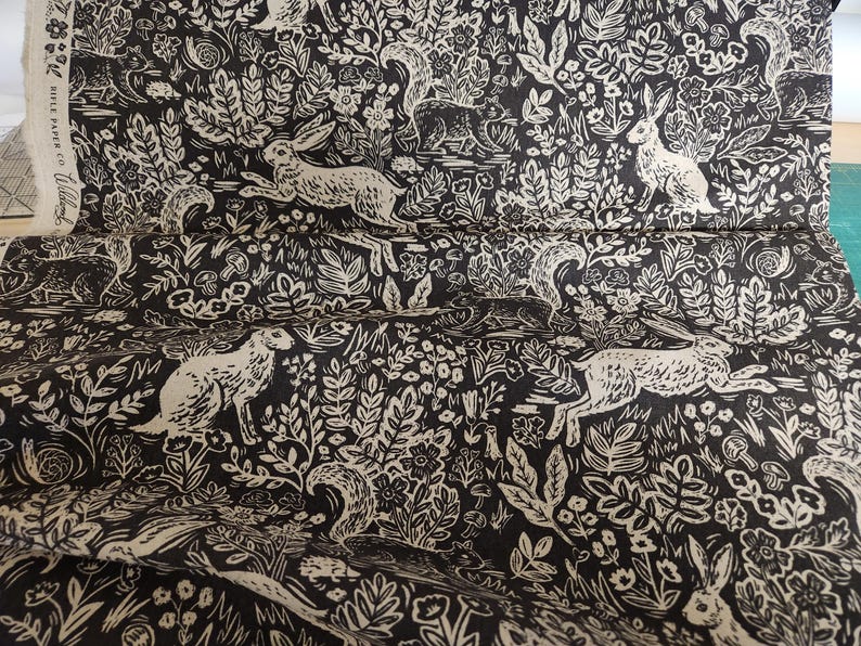 May include: Black and white fabric with a repeating pattern of stylized rabbits, squirrels, and floral designs. The fabric has a vintage, woodblock print aesthetic.
