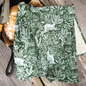May include: A green and white patterned kitchen towel with a woodland design featuring rabbits, squirrels, and foliage. The towel is folded over a cast iron skillet with a loaf of bread and a bread knife.