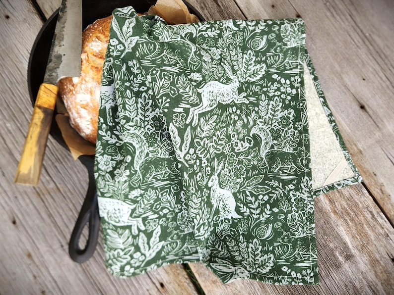 May include: A green and white patterned kitchen towel with a woodland design featuring rabbits, squirrels, and foliage. The towel is folded over a cast iron skillet with a loaf of bread and a bread knife.