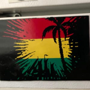 May include: A rectangular magnet with a red, yellow, and green design, featuring a palm tree silhouette. The magnet is affixed to a white surface, surrounded by other magnets and stickers.