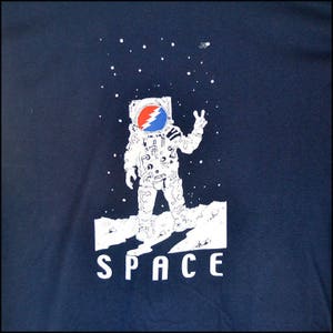 May include: Navy blue t-shirt featuring a white astronaut with a red, blue, and white logo on the helmet, giving a peace sign. The word "SPACE" is printed in white below the astronaut, with white stars in the background.