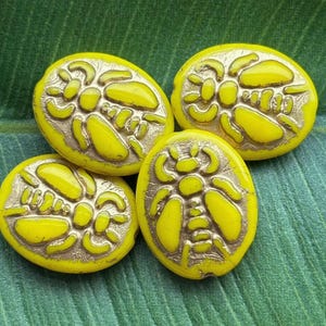 May include: Three oval-shaped beads featuring a raised beetle design. Each bead has a bright yellow surface with metallic gold-colored recessed details highlighting the insect's body, wings, and legs. The beads are arranged in a cluster.