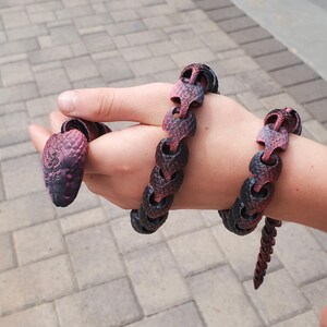 May include: A flexible, articulated snake toy featuring a textured, scale-like surface with a gradient color pattern transitioning from deep black to a vibrant reddish-purple. The segmented body allows for fluid movement and realistic coiling.