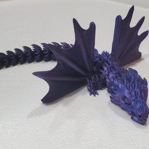 May include: A 3D printed articulated dragon figurine in a deep metallic purple color. The creature features a long, segmented tail, two large upright wings with pointed tips, and a textured body with scales covering the head and back.
