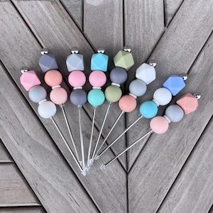 Peut inclure: Une collection de piques à cocktail décoratives. Chaque pique est composée d'une tige argentée surmontée d'une combinaison de perles géométriques et sphériques aux tons pastel de rose, gris, bleu et vert. Les piques sont disposées sur une surface en bois.