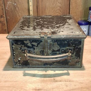 Puede incluir: Una caja metálica antigua con tapa con bisagras y asa. La caja es rectangular, con un acabado oscuro y desgastado. Una regla de madera se coloca en la parte superior de la caja.