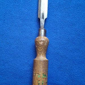 May include: A woodworking chisel with a wooden handle and a silver blade. The handle is a light brown color with a textured design. The blade is rectangular and has a sharp edge. The chisel is set against a blue background.