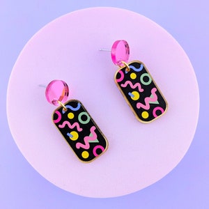 May include: A pair of geometric dangle earrings. Each earring has a black rectangular pendant with a gold border, decorated with colourful abstract shapes. A pink circular stud tops each earring, creating a vibrant contrast against the black background.