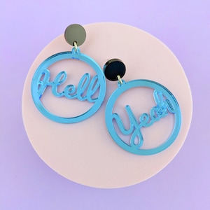 May include: A pair of blue acrylic earrings. Each earring features a circular design with the words "Hell" and "Yeah" cut out. The earrings have black and grey circular studs.