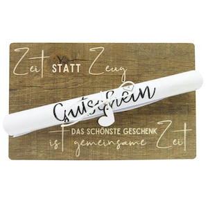 Può includere: Un cartello di legno con il testo "Zeit statt Zeug" e "Das schönste Geschenk ist gemeinsame Zeit" con un buono regalo bianco arrotolato che dice "Gutschein" legato con uno spago e un cuore.