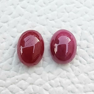 Könnte beinhalten: Zwei ovale, polierte Edelsteine. Einer ist tiefrot, der andere ein helleres, rötliches Violett. Beide haben eine glänzende, reflektierende Oberfläche, möglicherweise Cabochons. Die Edelsteine liegen auf einem weißen, strukturierten Hintergrund.