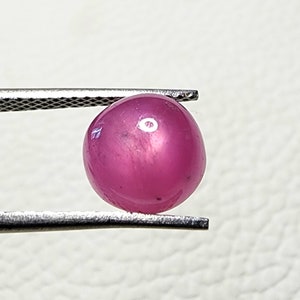 May include: A round, pink gemstone with a star-like reflection in the centre. The gemstone is held in a pair of tweezers.