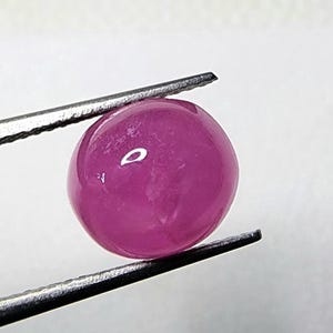 May include: A round, polished, magenta gemstone is held by a pair of silver tweezers. The gemstone has a smooth, reflective surface, and the tweezers are positioned to hold it securely. The background is a plain white.