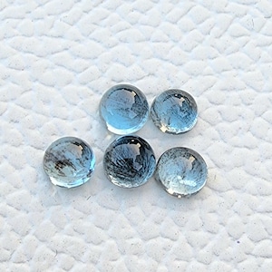 May include: Five round, light blue gemstones with a polished, reflective surface. The gems are arranged on a white, textured background. The stones have a slight internal pattern, adding depth to their appearance.