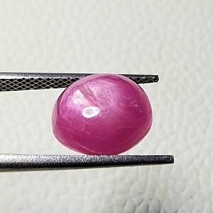 Puede incluir: Una piedra preciosa ovalada de color rosa con un patrón en forma de estrella en su interior. La piedra preciosa está sujeta con unas pinzas.