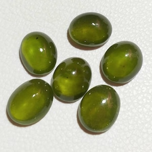 Op de afbeelding: Zes ovale, groene edelstenen met een gladde, gepolijste afwerking. De stenen zijn in een cluster op een wit oppervlak gerangschikt.