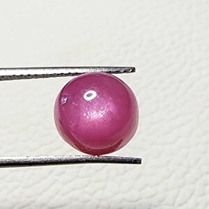 May include: A round, pink gemstone with a star-like pattern in the centre. The gemstone is held by a pair of tweezers.