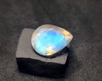 May include: A pearlescent teardrop-shaped gemstone with iridescent blue and white hues. The gemstone is displayed on a black velvet cushion.