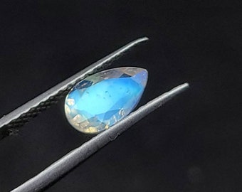 May include: A pear-shaped, iridescent gemstone with a blue and white colour gradient. The gem is held by silver tweezers against a black background. The gemstone's facets reflect light, creating a shimmering effect.