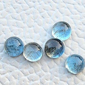May include: Five clear, round, blue glass marbles are arranged on a textured white surface. The marbles have a slight iridescence and reflect light, creating a shimmering effect. The marbles are a light blue color.