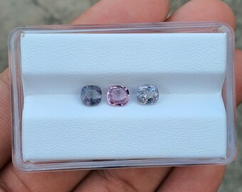 Pode incluir: Três pedras preciosas soltas em uma vitrine branca. As pedras preciosas são de cor roxo claro, rosa claro e azul claro.