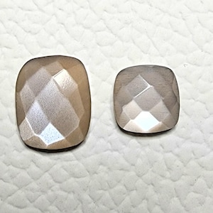 May include: Two faceted, square-cushion cut gemstones. The larger stone is a warm, light brown color, while the smaller stone is a lighter, greyish-brown. Both stones have a reflective, polished surface, and are set against a white textured background.