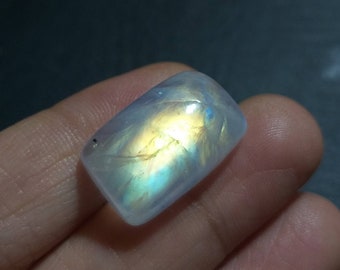 May include: A rectangular, polished moonstone gemstone with a rounded top. The stone displays a shimmering play of color, with hues of yellow, blue, and white. The gemstone is held in a hand.