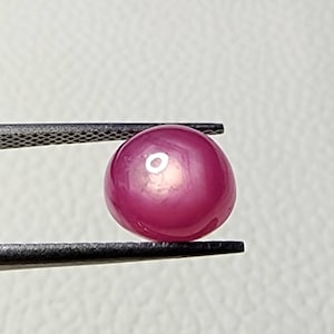 Könnte beinhalten: Ein runder, rosafarbener Edelstein mit einem sternförmigen Muster in der Mitte. Der Edelstein wird mit einer Pinzette gehalten.