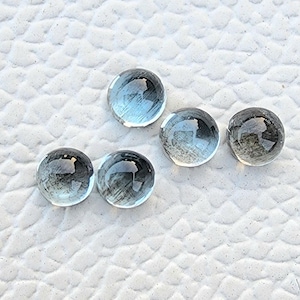 May include: Five round, translucent gemstones with a light blue hue. The gems are arranged on a textured, white surface. The stones have a polished, reflective appearance, suggesting they are cut and faceted.