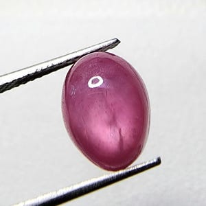 May include: A polished, oval-shaped, ruby gemstone held by tweezers. The gem is a deep pinkish-red color, with a smooth, reflective surface. The tweezers are silver-colored and hold the gemstone securely.