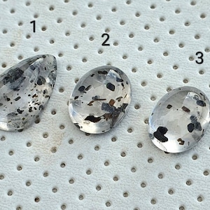 Puede incluir: Tres piedras de cuarzo transparentes con inclusiones negras, etiquetadas con 1, 2 y 3. Las piedras están sobre un fondo blanco con un patrón de pequeños agujeros.