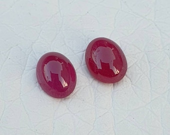 May include: Two oval-shaped, polished gemstones with a deep red hue. The gems have a smooth, glossy surface, reflecting light. The stones are likely rubies, often used in jewelry.