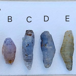 May include: Lot A: Six faceted, pointed, blue and clear sapphire crystals arranged in a row on a white background. The crystals are labeled A through F.