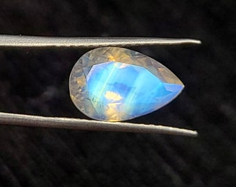 May include: A pear-shaped, faceted moonstone gemstone held by tweezers. The stone displays a captivating play of colour, with iridescent blue and yellow hues. The gemstone is set against a dark background, highlighting its brilliance.