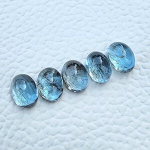 May include: Five oval-shaped, light blue gemstones are arranged diagonally across a white, textured surface. The gemstones have a translucent quality, with internal patterns and variations in color, ranging from pale to medium blue.