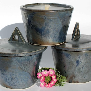 Könnte beinhalten: Drei Kerzenhalter aus Keramik mit Deckeln. Die Halter sind dunkelblau mit einer marmorierten Glasur. Die Deckel haben einen dreieckigen Griff mit einem Loch in der Mitte. Die Kerzenhalter sind übereinander gestapelt.