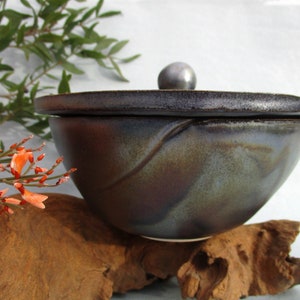 Könnte beinhalten: Eine dunkelbraune Keramikschüssel mit Deckel. Die Schüssel hat eine gefleckte Glasur in Blautönen, Braun- und Schwarztönen. Der Deckel hat einen kleinen, runden Knopf oben.