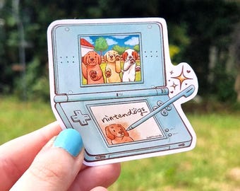 May include: A light blue handheld gaming console sticker featuring a dual-screen display. The top screen shows three cartoon puppies, while the bottom screen displays the text nintendogs with a puppy illustration and a stylus resting on the surface.