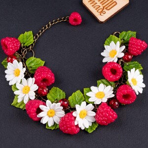 Op de afbeelding: Een handgemaakte armband met witte madeliefjes en rode frambozen, allemaal gemaakt van polymer clay. De armband is gemaakt met een bronzen ketting en heeft een handgeschilderd "Hand Made"-label.