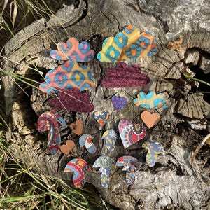 May include: Assortment of fabric cutouts in various shapes and patterns, including clouds, hearts, and mushrooms. The cutouts are arranged on a wooden surface with a natural bark texture.