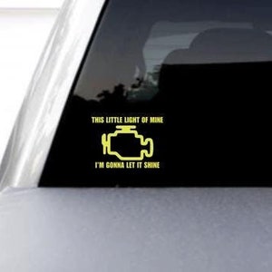 May include: A yellow check engine light sticker on the rear window of a silver truck. The sticker says "This little light of mine, I'm gonna let it shine."