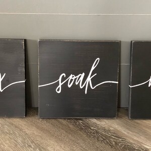 May include: Three black wooden signs with white lettering. The signs read "relax", "soak", and "unwind".