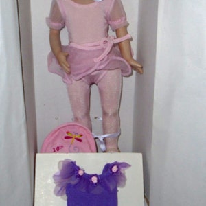 May include: A red-haired doll wearing a pink leotard and tutu with a white ribbon around her neck. The doll is holding a pink and white hat with a dragonfly design and the text "10th" on it. The doll is standing on a white surface with a purple leotard and tutu on a white card.